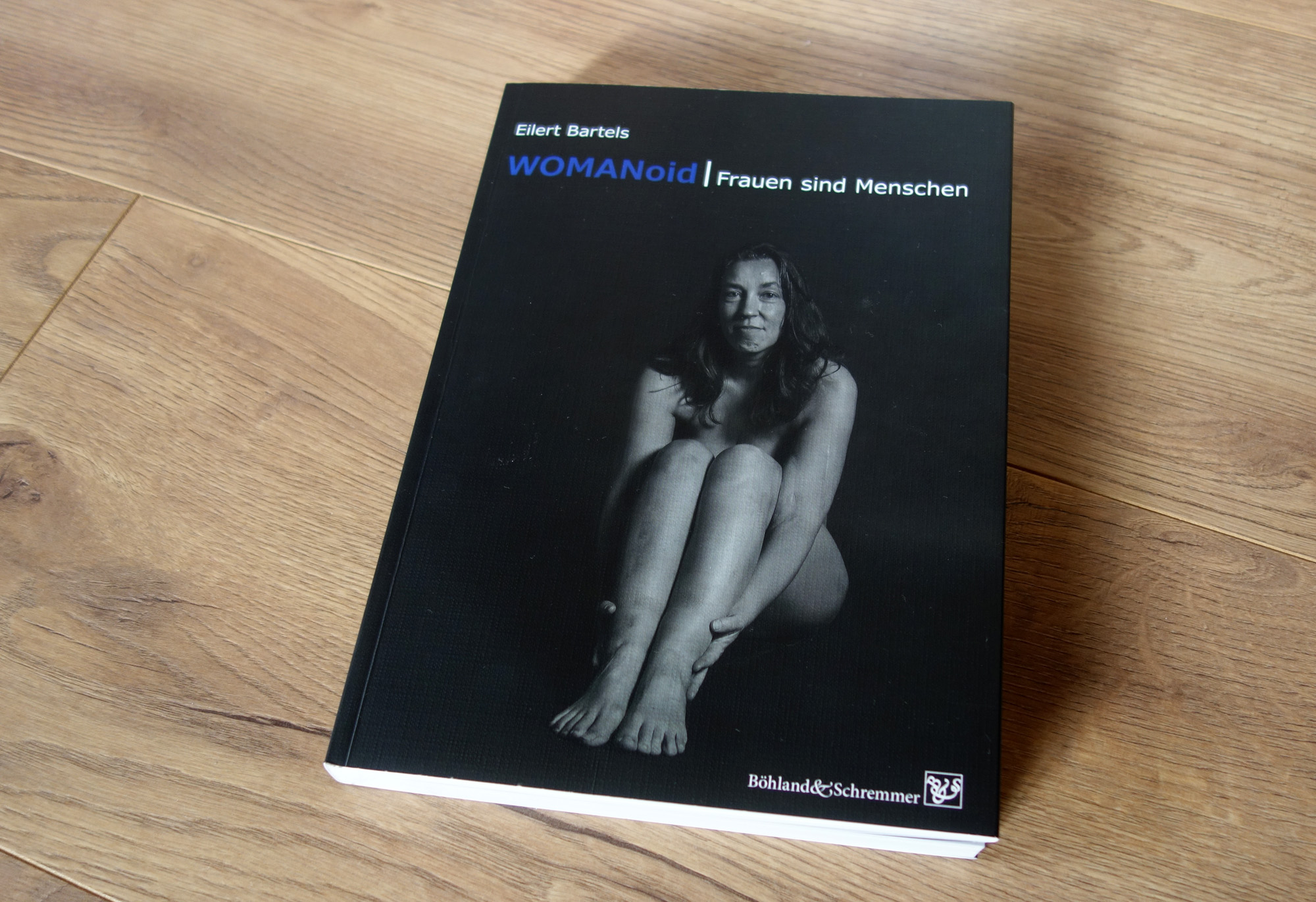 Das Buch WOMANoid fotografiert auf dem Boden liegend. Auf dem Cover sitzt eine nackte Frau mit angezogenen Beinen und lächelt verhalten in die Kamera.
