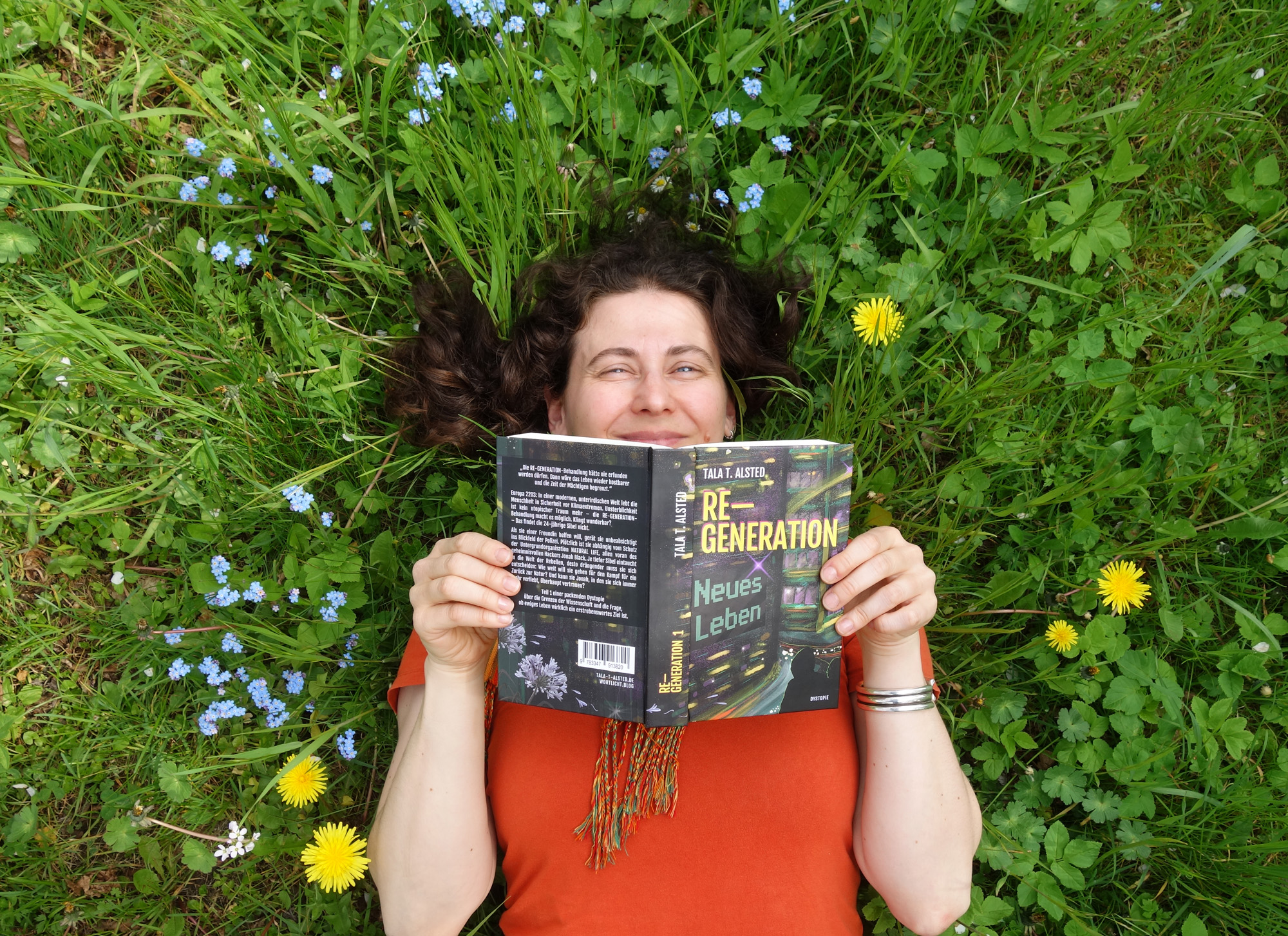 Tala T. Alsted mit ihrem Buch "RE-GENERATION - Neues Leben" auf einer Wiese mit Löwenzahn- und Vergissmeinnicht-Blüten